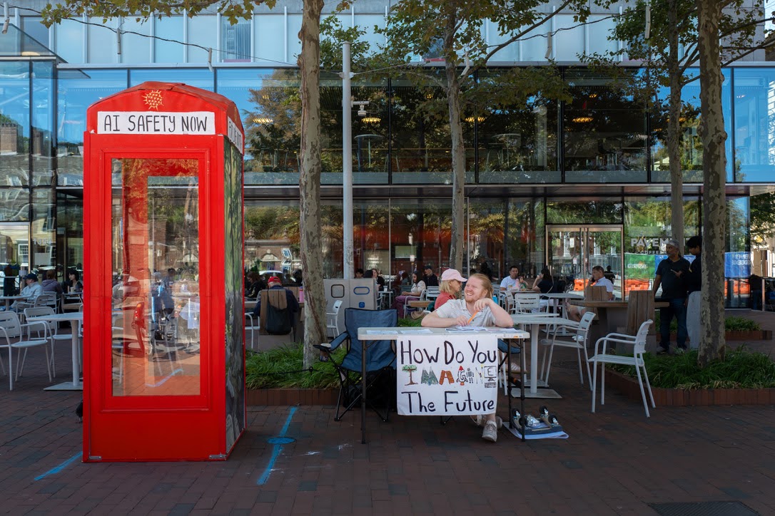 A red AI Safety Now phone booth beside a community outreach table
