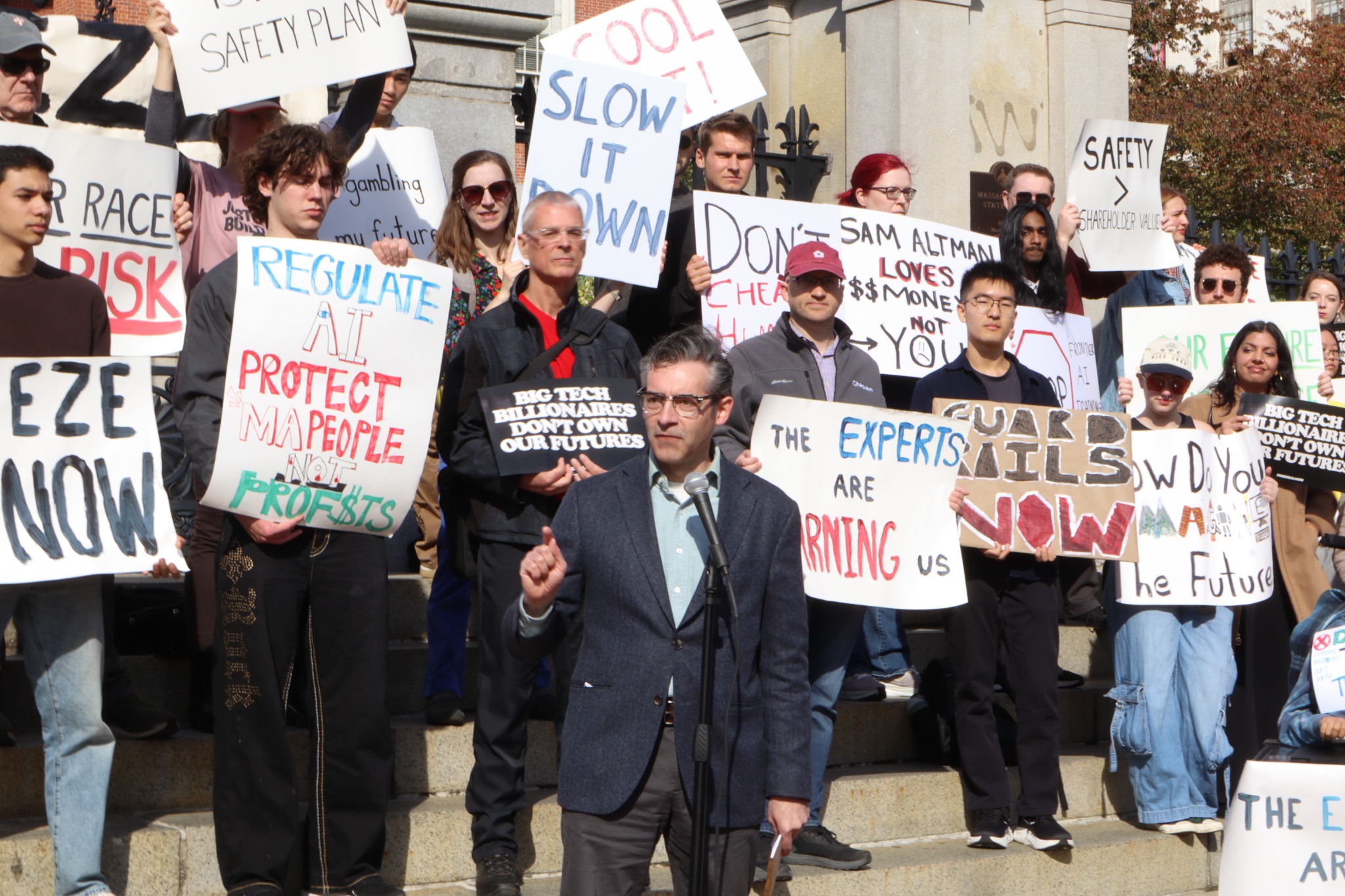 A speaker addressing a rally outside the Massachusetts State House, with people holding AI safety signs